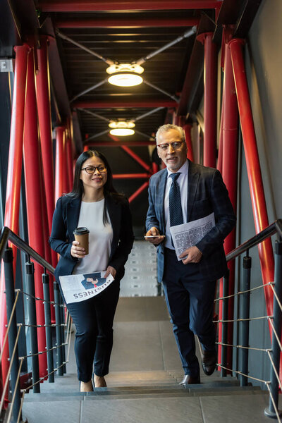 улыбающийся бизнесмен и азиатская бизнесвумен с газетами, смотрящими в камеру
 