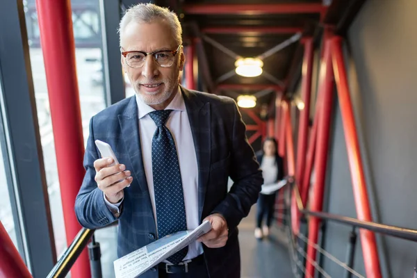 Leende Affärsman Kostym Med Tidning Och Smartphone Tittar Kameran — Stockfoto