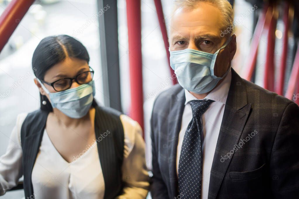 selective focus of businessman and asian businesswoman in medical masks looking at camera 