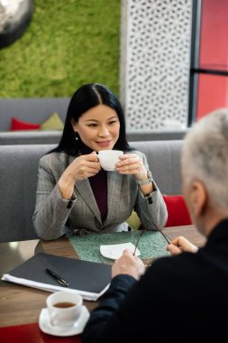 gülümseyen Asyalı iş kadınının seçici odak noktası iş adamıyla konuşması ve kahve bardağını tutması. 