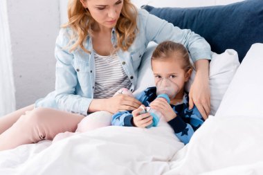caring mother sitting near sick daughter using inhaler with spacer  clipart