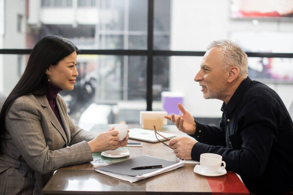 side view of businessman and smiling asian businesswoman talking in cafe 