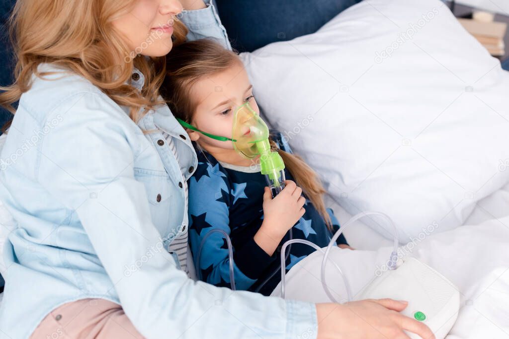 mother near asthmatic kid in respiratory mask using compressor inhaler in bedroom 