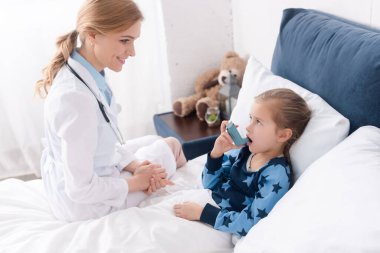 Mutlu doktor astımlı çocuğa bakıyor ağzı açık bir şekilde astım spreyi kullanıyor. 