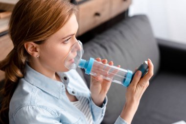 asthmatic woman using inhaler with spacer in living room  clipart