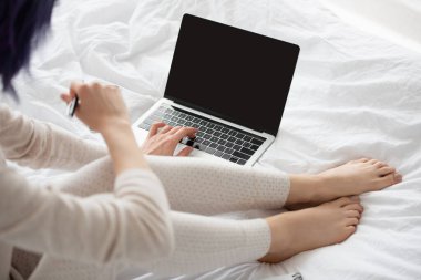 Cropped view of freelancer with pen working on laptop on bed clipart