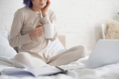 Cropped view of freelancer with crossed legs holding cup of tea near laptop on bed clipart