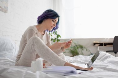 Renkli saçlı, dizüstü bilgisayarın yanında akıllı telefon ve yatakta çay kullanan serbest çalışan biri.