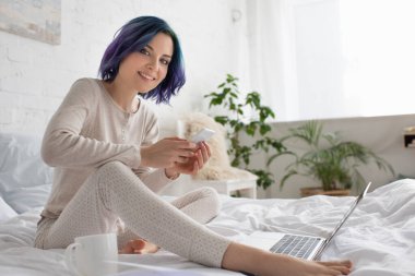 Renkli saçlı serbest çalışan, gülümseyen, kameraya bakan, bilgisayarın yanında akıllı telefon kullanan ve yatakta çay içen biri.