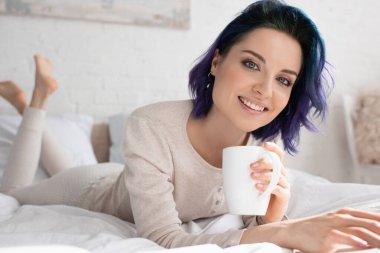 Renkli saçı ve çay fincanı olan serbest çalışanın seçici odağı kameraya bakıyor, gülümsüyor ve yatakta uzanıyor.