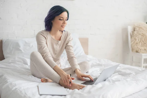 Freelancer with colorful hair holding pen and working on laptop near copybook on bed