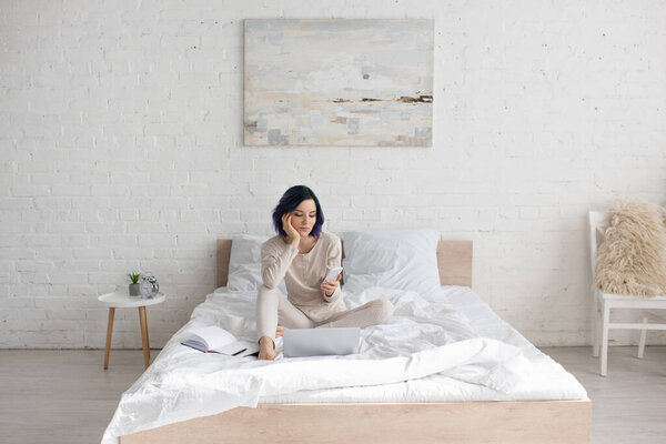 Freelancer with colorful hair using smartphone near laptop and notebook on bed in bedroom