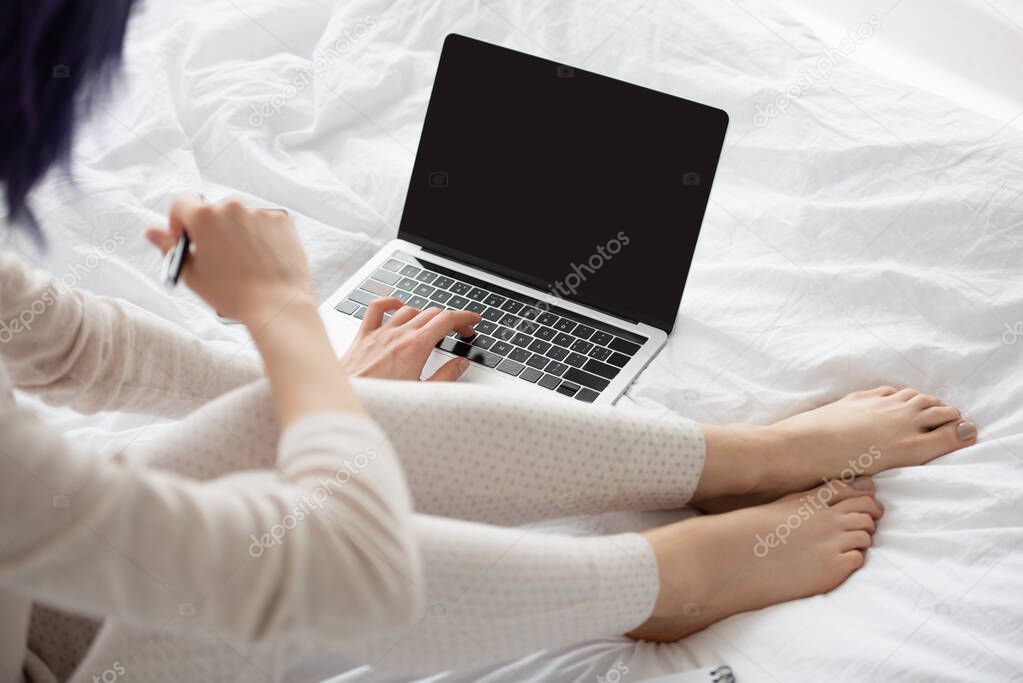 Cropped view of freelancer with pen working on laptop on bed