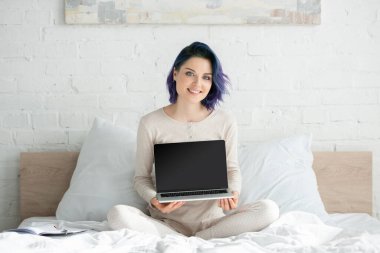 Freelancer with colorful hair and crossed legs showing laptop, smiling and looking at camera on bed in bedroom clipart