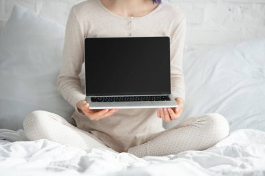 Cropped view of freelancer with colorful hair and crossed legs showing laptop on bed in bedroom clipart