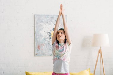 Renkli saçlı ve elleri sıkılmış bir kadın oturma odasında yoga yapıyor.
