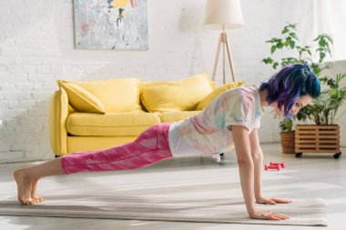Renkli saçlı bir kadın oturma odasında yoga minderinde tahta yapıyor.