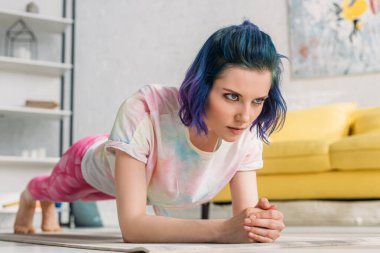 Selective focus of tense girl with colorful hair doing plank on yoga mat in living room clipart