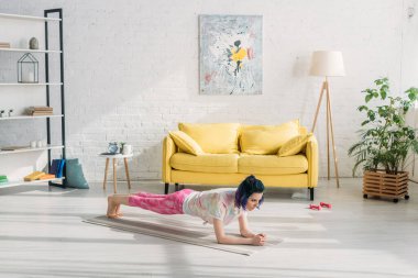 Renkli saçlı kız oturma odasındaki kanepenin yanındaki yoga minderinde kalas yapıyor.