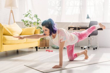 Renkli saçlı kadın, uzanmış elleriyle Asana 'yı yapıyor ve oturma odasında yoga minderinin üstünde bacağını kaldırıyor.