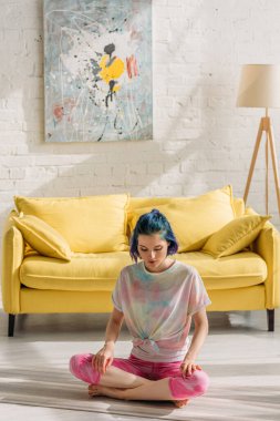 Renkli saçlı, bacak bacak üstüne atmış yoga minderinde oturmuş, oturma odasında meditasyon yapan bir kız.