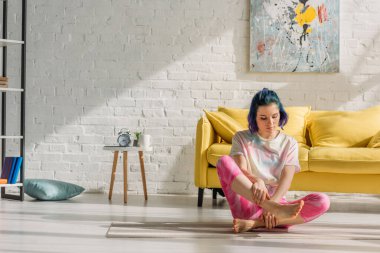 Renkli saçlı kız oturma odasında yoga minderinde bacağına dokunuyor.