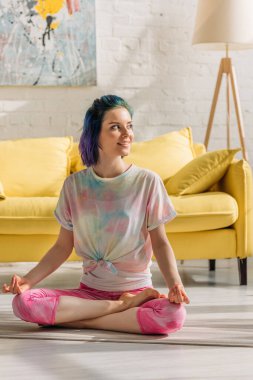 Renkli saçlı, gülümseyen, bakışlarını kaçıran ve oturma odasında yoga minderinde meditasyon yapan bir kız.