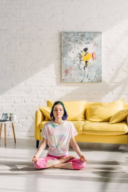 Renkli saçlı ve gözleri kapalı bir kız oturma odasında yoga minderi üzerinde meditasyon yapıyor.