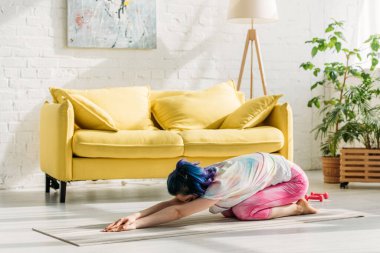 Çocukluğunda renkli saçlı bir kız oturma odasında yoga minderinde poz veriyor.