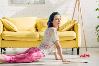 Kobralı renkli saçlı kız yoga minderinde poz verip oturma odasındaki kameraya bakıyor.