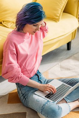 Renkli saçlı, çapraz bacaklı serbest çalışan, kanepenin yanında dizüstü bilgisayarla çalışan biri.