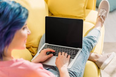 Selective focus of freelancer with colorful hair and laptop smiling and lying on sofa clipart