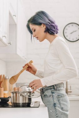 Tava kapağı ve sobanın yanında spatula tutan renkli saçlı bir kız.