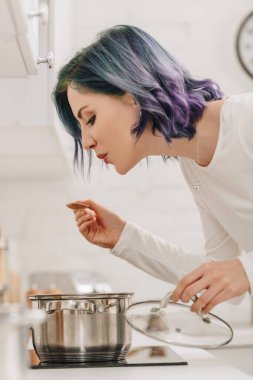 Tava kapağı tutan ve mutfak sobası yanında spatula üfleyen renkli saçlı kızın seçici odağı.