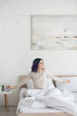 Renkli saçlı ve kolları açık çekici bir kadın yatak odasına bakıyor.