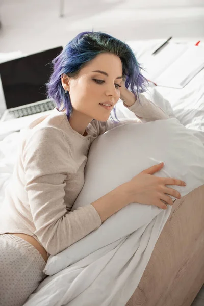 Dreamy freelancer with colorful hair lying near laptop on bed