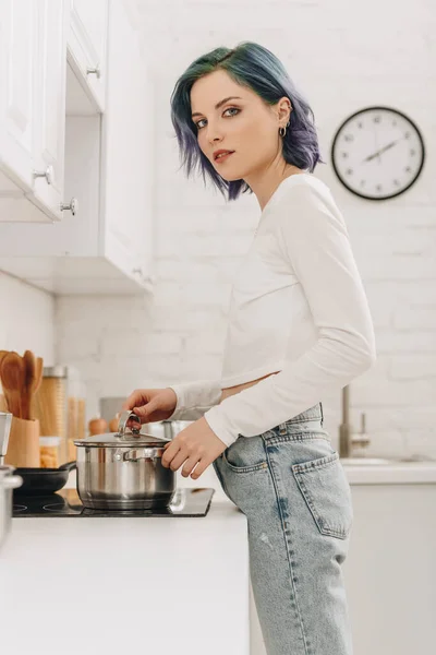 Low angle view of of girl with colorful hair touching pan lid near kitchen stove and looking at camera — Stock Image Low Angle View Girl Colorful Hair Touching Pan Lid Kitchen Stock Photo