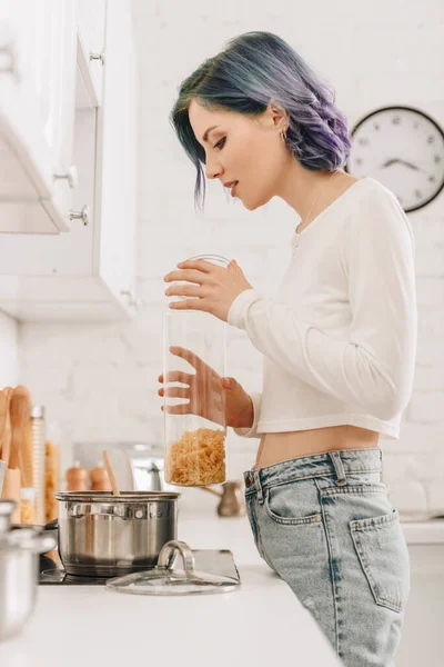 Selective focus of girl with colorful hair holding container with pasta near kitchen stove — Stock Image Selective Focus Girl Colorful Hair Holding Container Pasta Kitchen Stove Stock Image