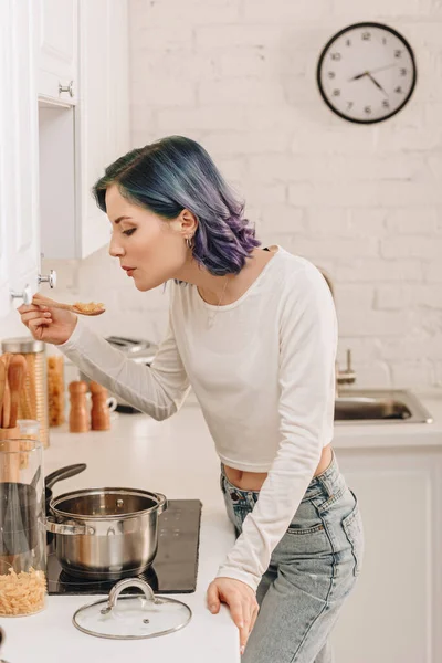 Girl with colorful hair blowing on spatula with pasta near kitchen stove — Stock Image Girl Colorful Hair Blowing Spatula Pasta Kitchen Stove Stock Picture