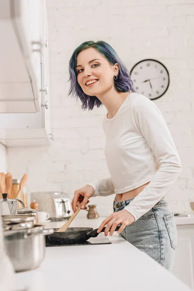 Low angle view of girl with colorful hair preparing food with spatula on frying pan, smiling and looking at camera in kitchen — Stock Image Low Angle View Girl Colorful Hair Preparing Food Spatula Frying Stock Image