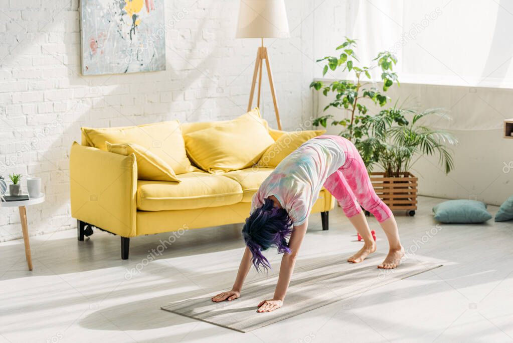 Woman with colorful hair in downward dog pose on yoga mat in living room