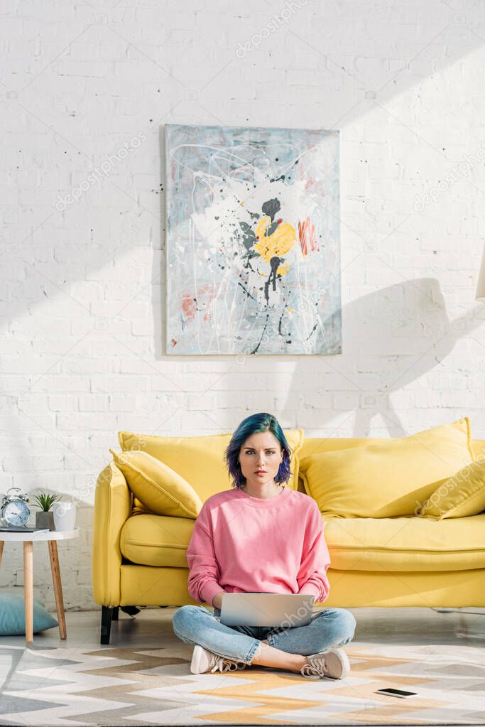 Freelancer with colorful hair and crossed legs looking at camera and working with laptop near sofa and smartphone on floor 