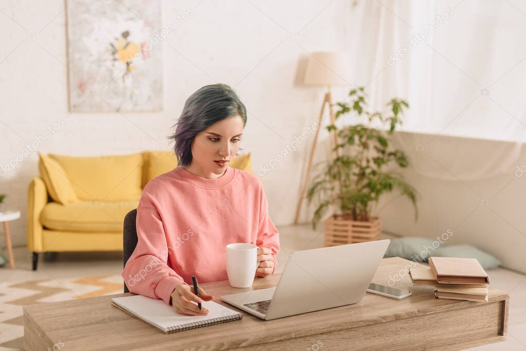 Freelancer with colorful hair holding cup of tea and pen above copybook near laptop at table 