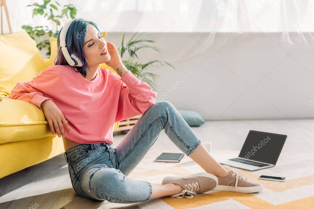 Freelancer with colorful hair, closed eyes and headphones listening music near sofa, laptop and smartphone on floor