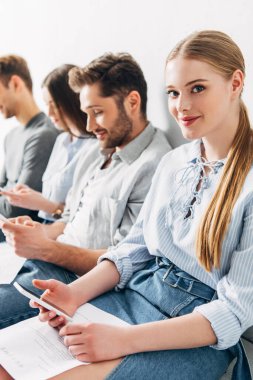Akıllı telefonlu gülümseyen kızın seçmeli odak noktası ve ofisteki adayların yakınındaki kameraya bakmak. 