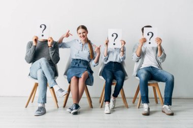 Cheerful girl pointing on oneself near employees holding cards with question marks in office  clipart