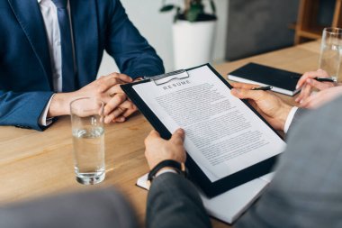 Cropped view of recruiter holding clipboard with resume near colleagues and employee  clipart