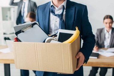 Selective focus of dismissed man holding cardboard box with papers, digital tablet and banana in office  clipart