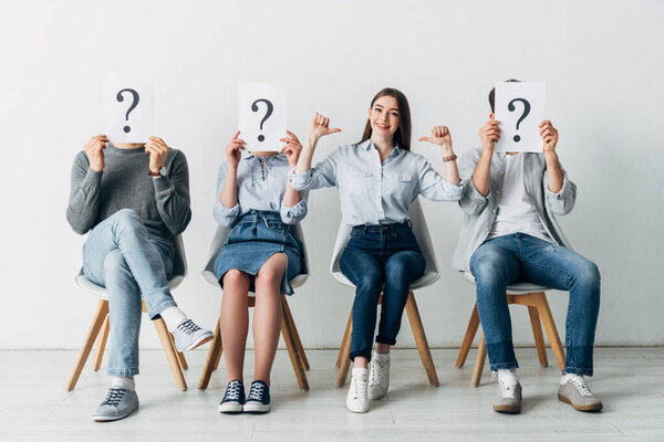 Улыбающаяся женщина показывает большие пальцы вверх рядом с сотрудниками с вопросительными знаками на карточках в офисе
 