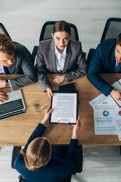 Overhead view of recruiter looking at camera near colleagues and employee with resume at table